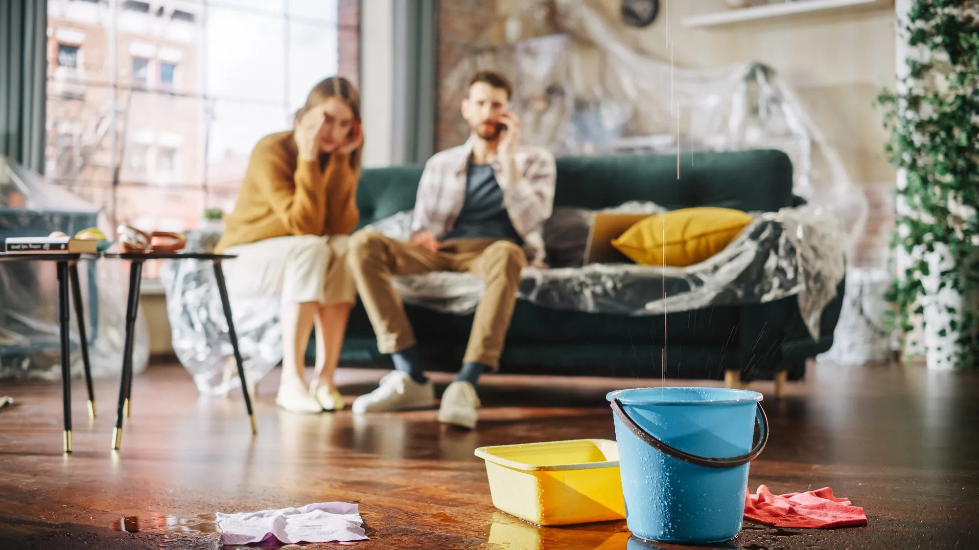 Water drips into a bucket and tray on a living-room floor while two people sit on a plastic-covered sofa, one on the phone.