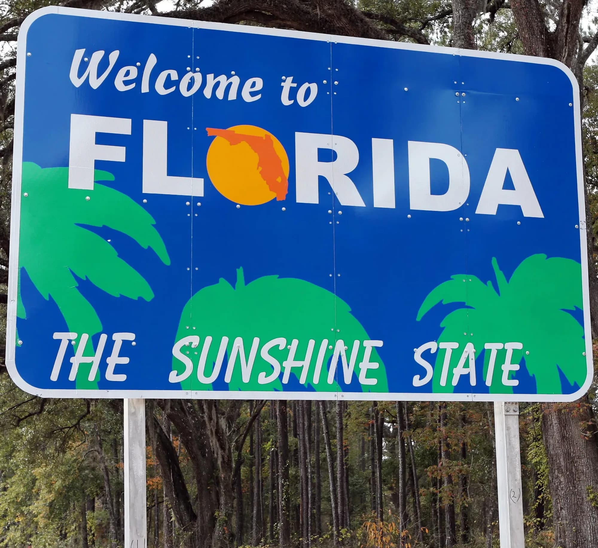 A roadside sign reading Welcome to Florida, The Sunshine State, with palm graphics and live oaks behind it.