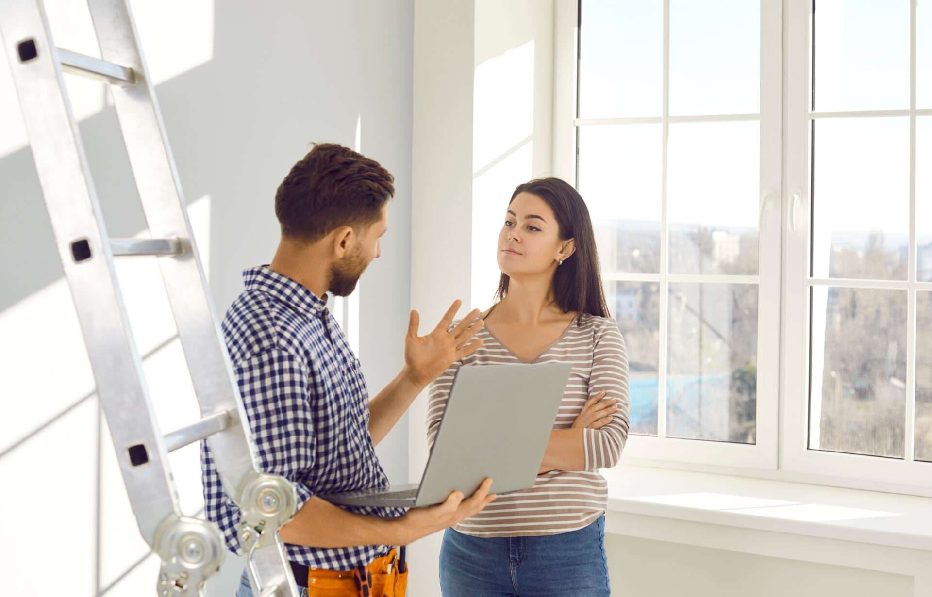 A contractor holding a laptop talks with a homeowner in an empty room, a stepladder beside them.