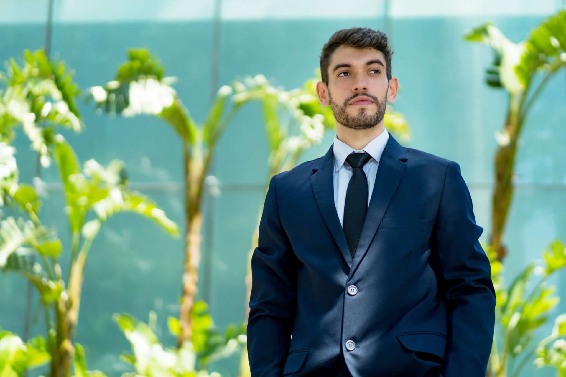 A man in a dark suit and tie standing outside a glass building, framed by tropical foliage.