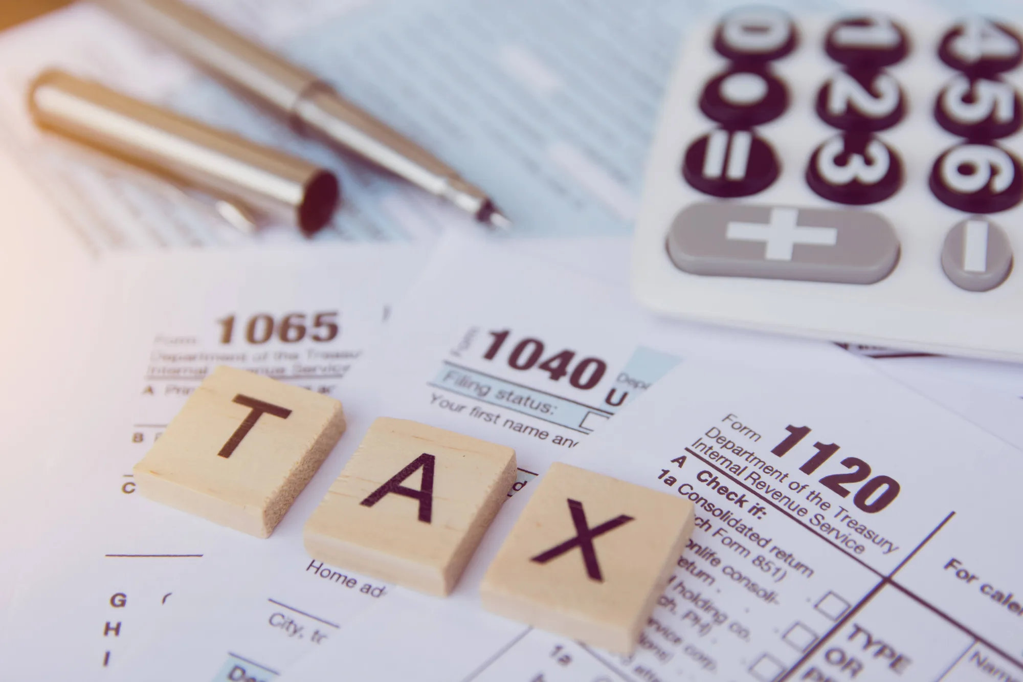 Wooden letter tiles spelling TAX resting on IRS forms 1040, 1065, and 1120 beside a calculator and pen.