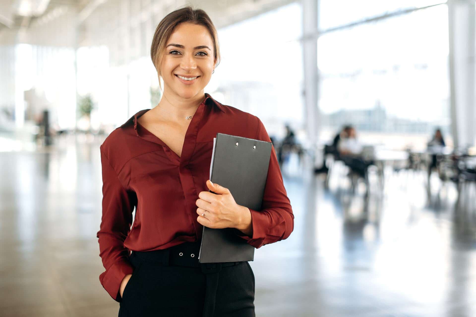 A woman in a dark red blouse holding a clipboard, smiling, in a bright open office.
