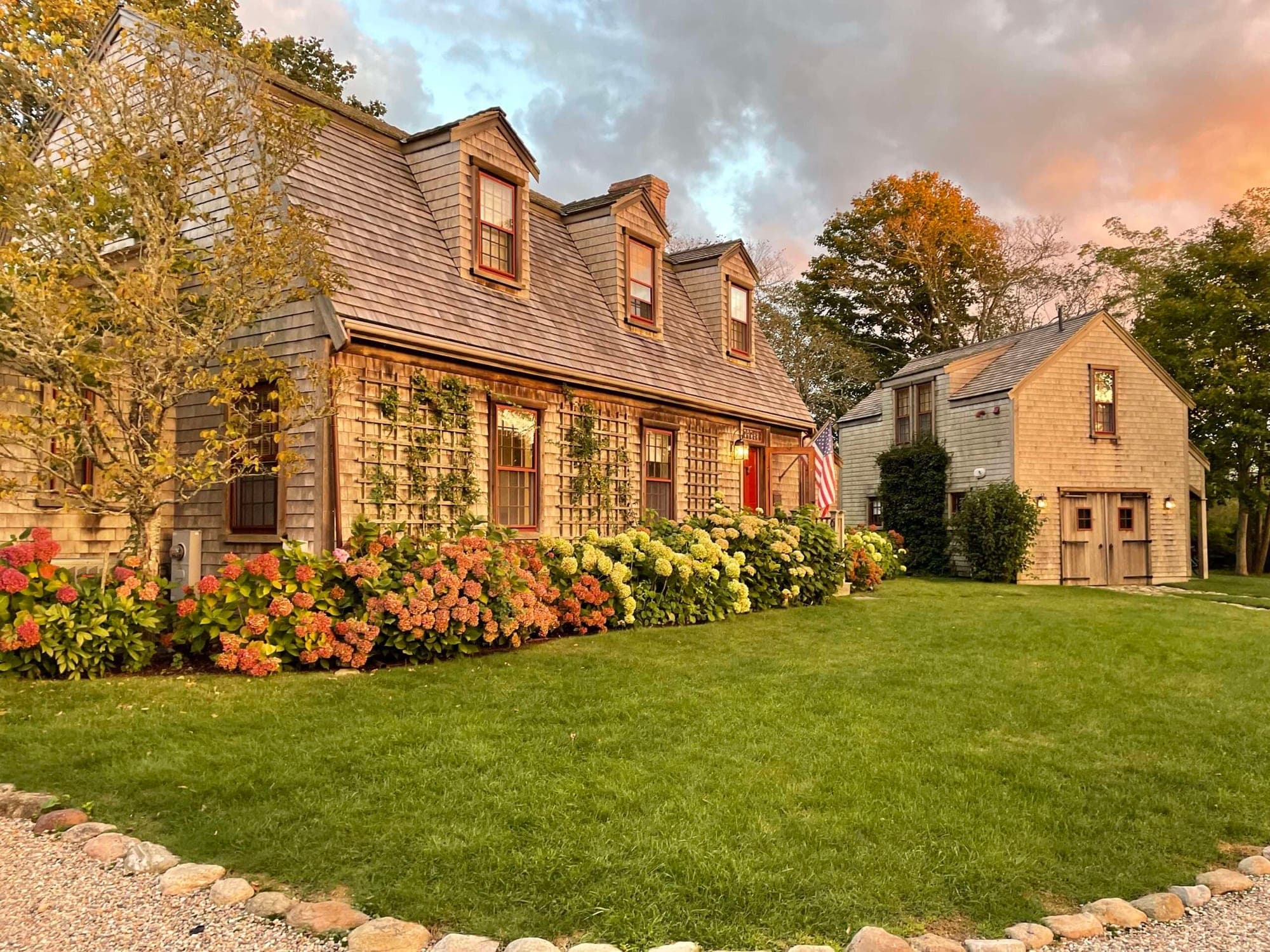 A cedar-shingled New England house with dormer windows and hydrangeas along the front, photographed at dusk.