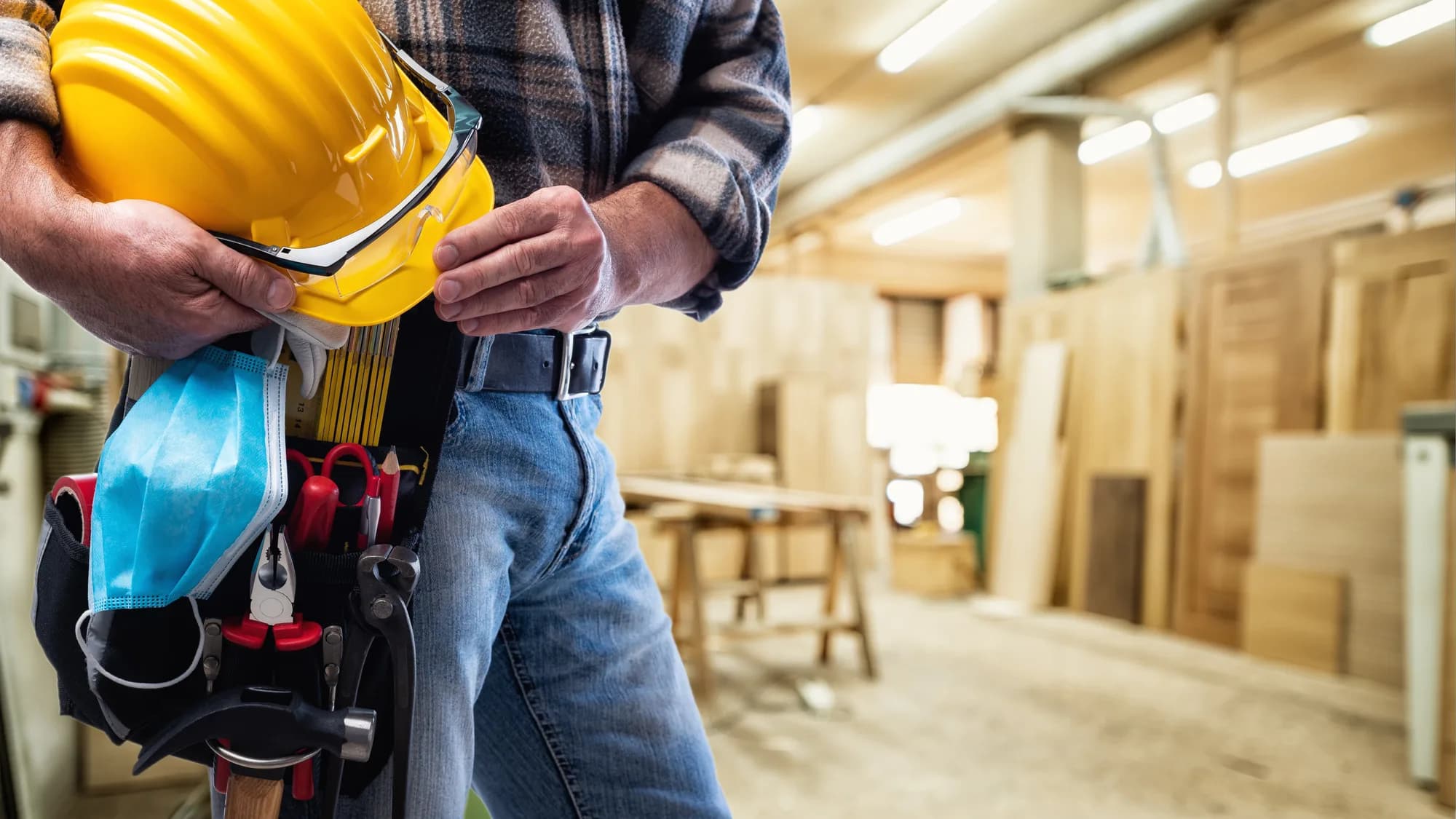 A tradesperson in a plaid shirt holding a hard hat and safety glasses, a tool belt at their waist, in a woodworking shop.